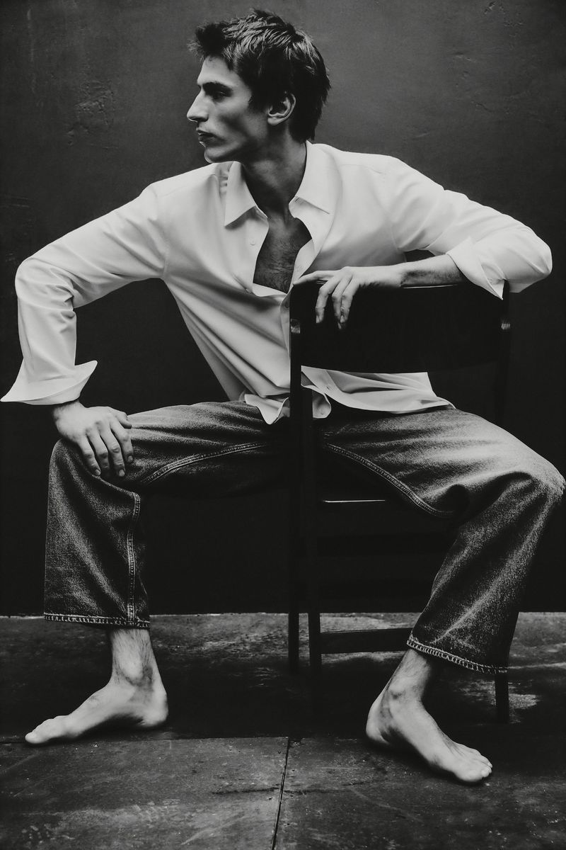 Monochrome portrait of a barefoot man seated backward on a dark chair wearing an open white button-down shirt and denim jeans.