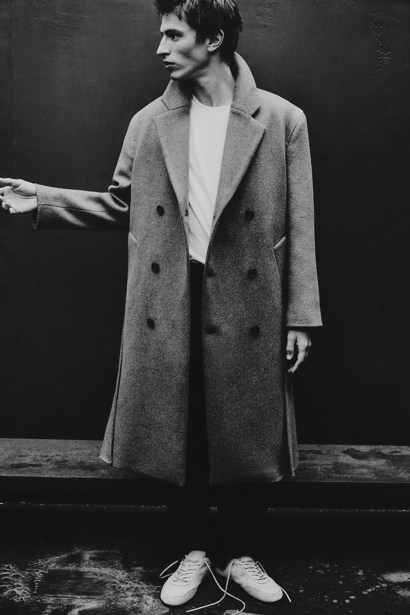 Black and white fashion shot of a slender adult man in an overcoat, t-shirt, and sneakers, standing against a dark backdrop.