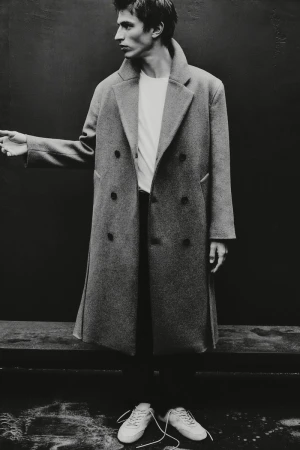 Black and white fashion shot of a slender adult man in an overcoat, t-shirt, and sneakers, standing against a dark backdrop.