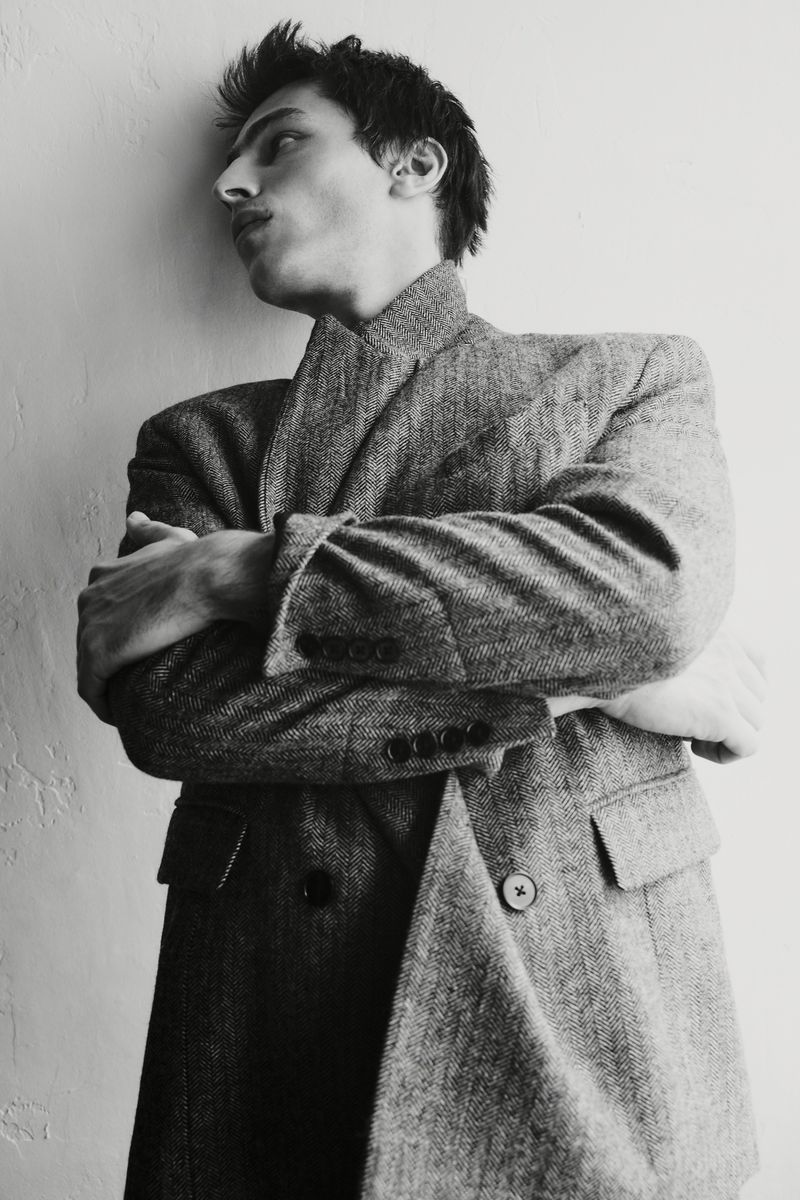 Portrait of a young adult male in a patterned grey overcoat posing against a white wall with arms crossed in a photo studio.