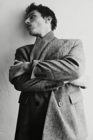 Portrait of a young adult male in a patterned grey overcoat posing against a white wall with arms crossed in a photo studio.