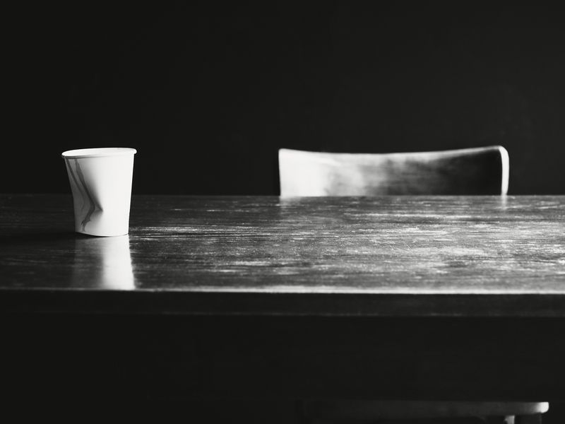 Paper cup sits on dark wooden table with chair in background against dark backdrop, minimal and somber.