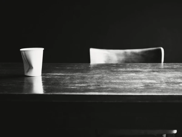 Paper cup sits on dark wooden table with chair in background against dark backdrop, minimal and somber.