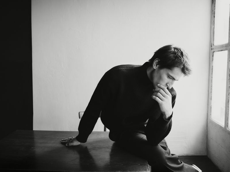 Monochrome portrait of a man seated on a dark table next to a window, wearing a black ribbed sweater.