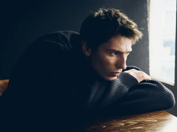 Model Clément Laguardia wearing a dark ribbed knit sweater, leaning on a wooden table near a bright window.