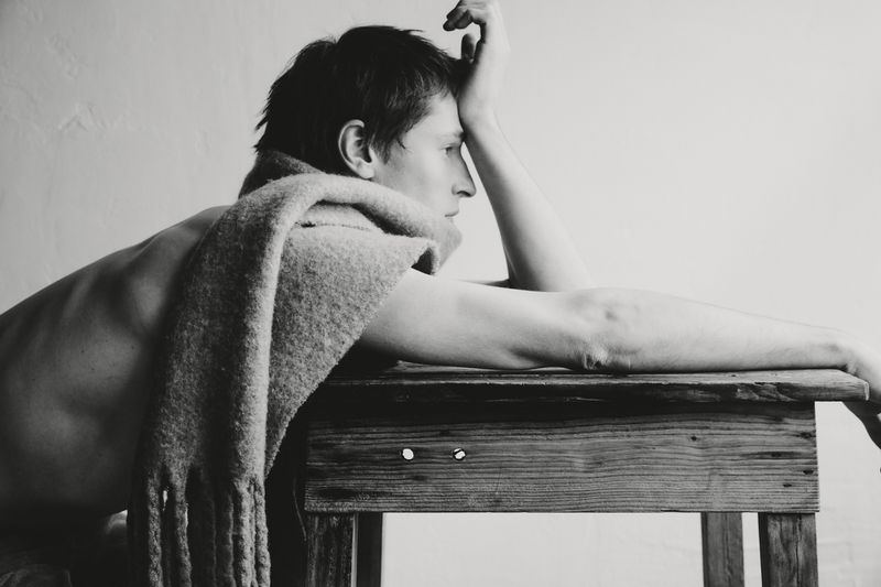 Monochrome portrait of a shirtless adult male with a scarf, resting on a wooden table against a plain white background.