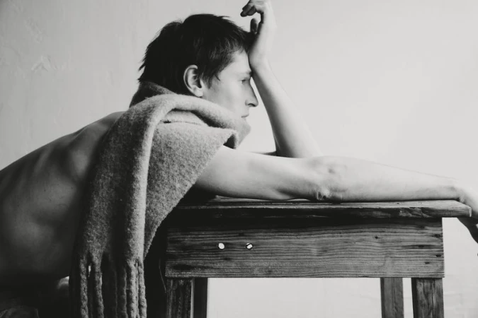 Monochrome portrait of a shirtless adult male with a scarf, resting on a wooden table against a plain white background.