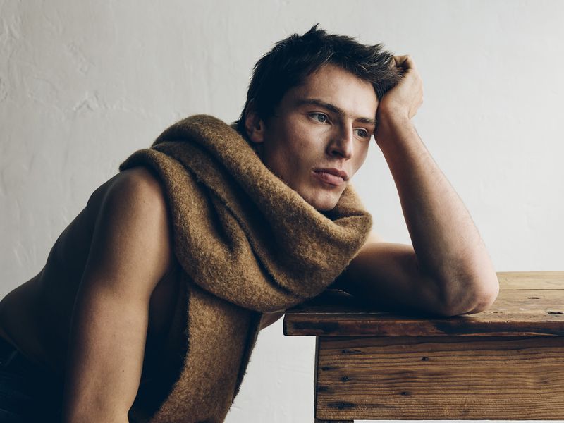 Portrait of a young adult male with a scarf draped around his neck, resting his head on a wooden table against a textured white background.