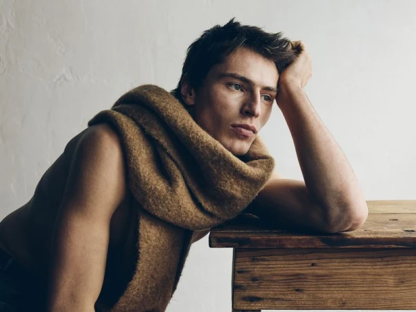 Portrait of a young adult male with a scarf draped around his neck, resting his head on a wooden table against a textured white background.