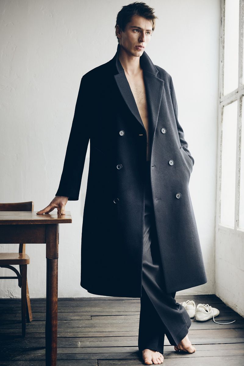 Male fashion model poses indoors wearing a long, dark overcoat and trousers, standing next to a wooden table and chair near a window.