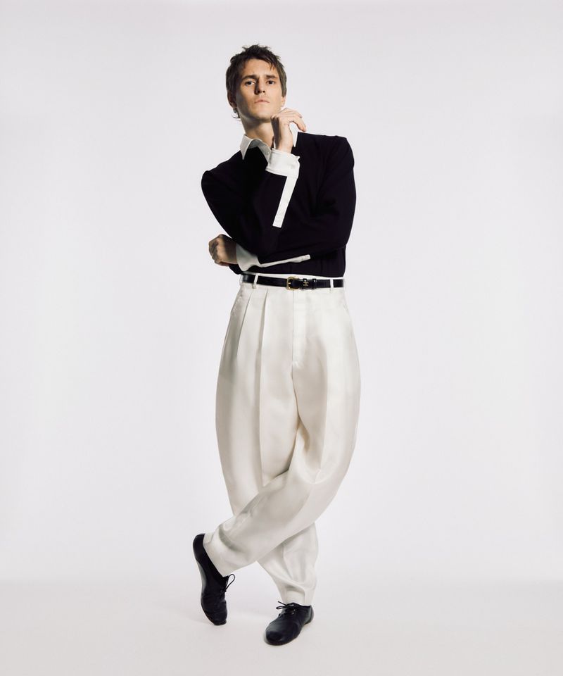 Full shot of a young adult male in a dark blazer, white dress shirt, pleated white slacks, and black shoes, posing in a photo studio.