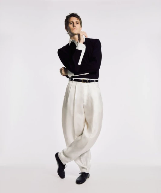 Full shot of a young adult male in a dark blazer, white dress shirt, pleated white slacks, and black shoes, posing in a photo studio.