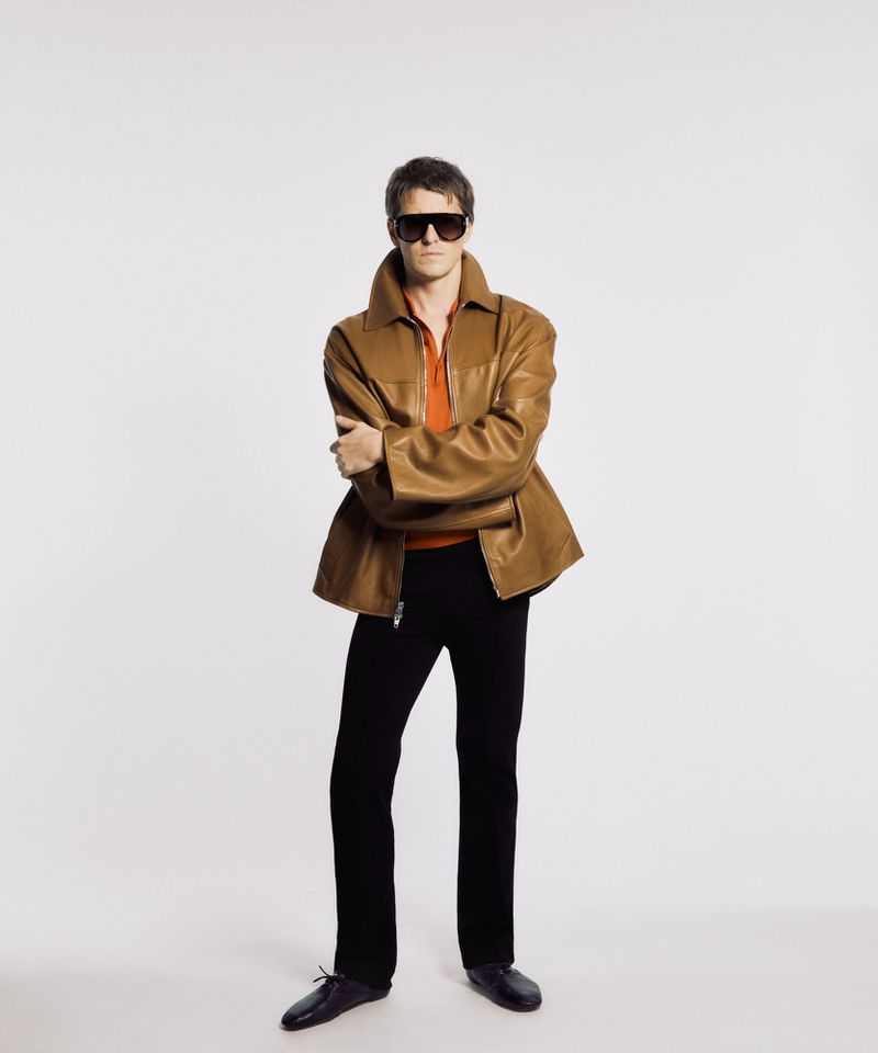 Full-length shot of an adult male posing in a brown jacket, orange shirt, black pants, and sunglasses against a white backdrop.