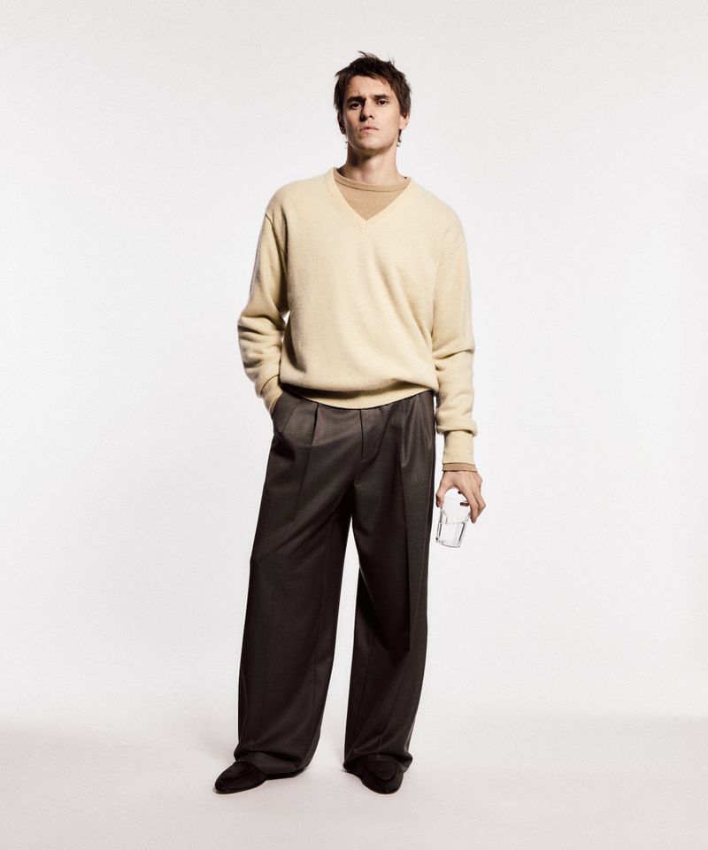 Full-length studio shot of a young adult male in a sweater and wide-leg pants holding a glass in neutral studio lighting.