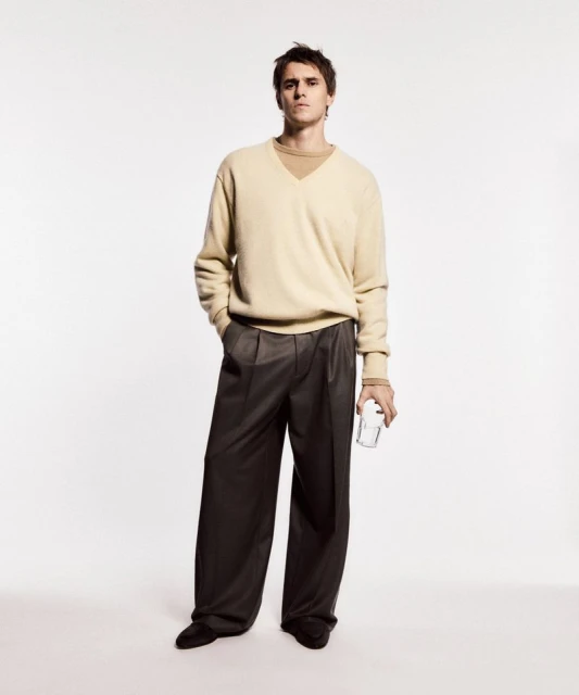 Full-length studio shot of a young adult male in a sweater and wide-leg pants holding a glass in neutral studio lighting.