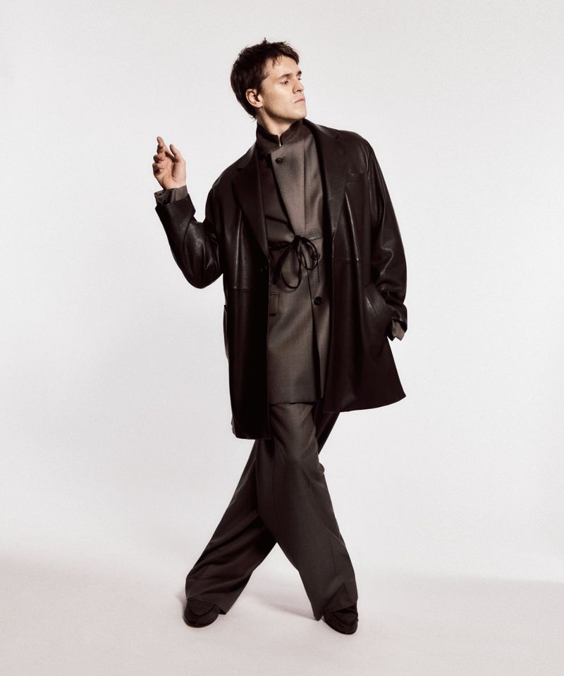 A young adult man in a dark coat and pants poses against a white background in a studio, showcasing modern fashion and style.