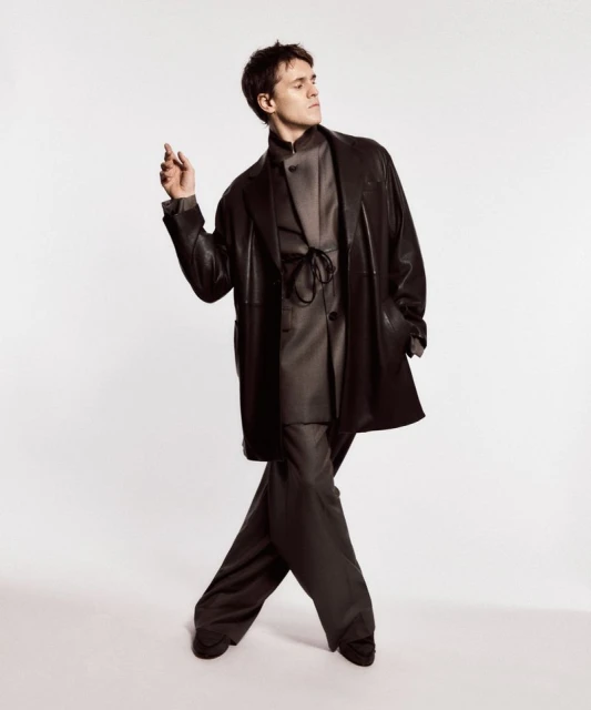 A young adult man in a dark coat and pants poses against a white background in a studio, showcasing modern fashion and style.