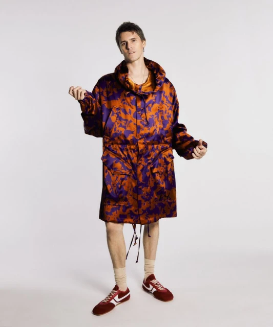 A young adult male stands in a studio wearing a long, patterned orange and purple parka with maroon running shoes against white.