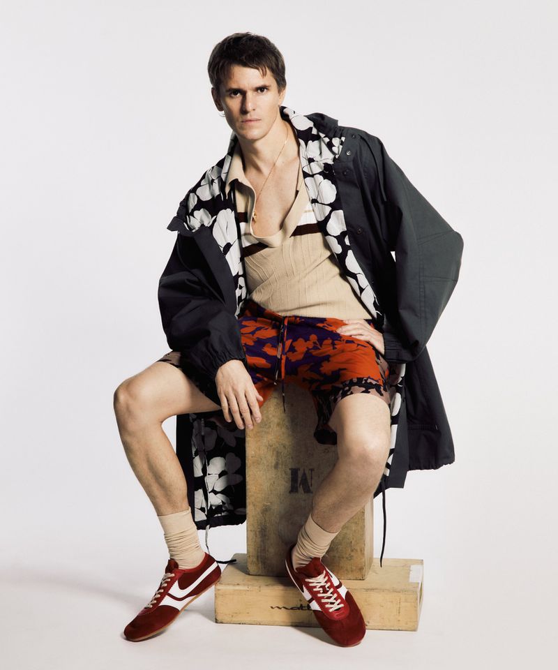 Male fashion model in an unbuttoned floral-print shirt, dark jacket, print shorts, tube socks, and red sneakers sitting in a photo studio.
