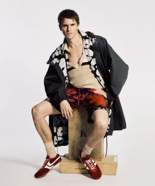 Male fashion model in an unbuttoned floral-print shirt, dark jacket, print shorts, tube socks, and red sneakers sitting in a photo studio.