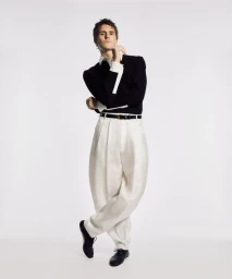 Model Adrien Dantou wearing pleated white trousers and a black long-sleeve knit top against a plain studio backdrop.
