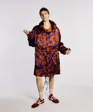 Model Adrien Dantou standing against a white backdrop in an oversized purple and bronze patterned hooded coat with red sneakers.