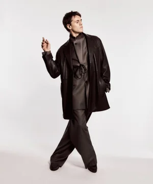 Model Adrien Dantou posing in a black leather coat and charcoal suit against a white studio background.