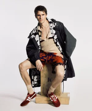 Male model seated on a wooden block wearing patterned shorts, beige polo shirt, dark jacket, and red sneakers against a white background
