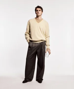 Model Adrien Dantou poses in a cream knit v-neck sweater and charcoal wide-leg trousers while holding a glass on a white backdrop.