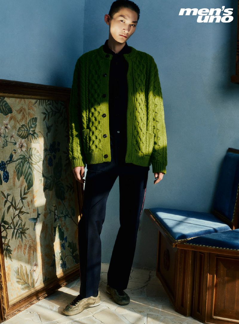 A young Asian man in a green cardigan poses indoors near vintage decor. The photo features muted colors and natural light, styled for a fashion editorial.
