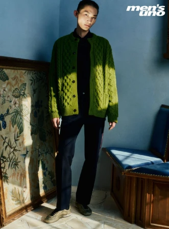 A young Asian man in a green cardigan poses indoors near vintage decor. The photo features muted colors and natural light, styled for a fashion editorial.