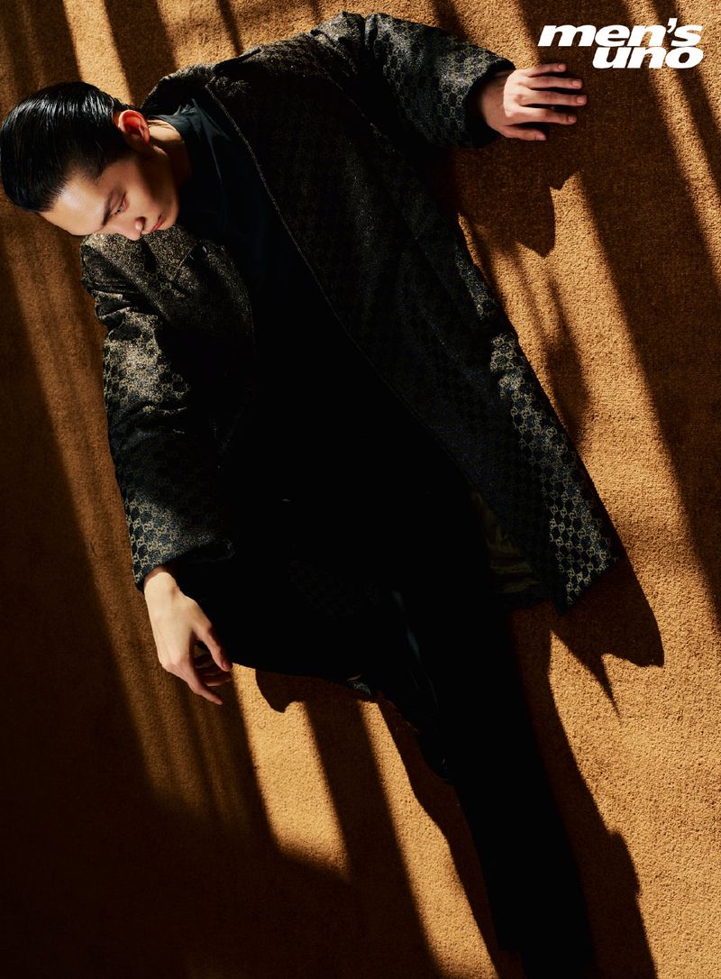 An adult male laying on a tan surface with shadows and bright light wearing a patterned black coat and dark pants, Men's Uno magazine logo visible.