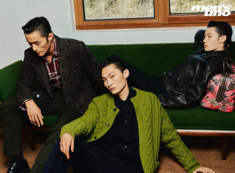 Three Asian male fashion models pose on a green velvet sofa in a studio wearing sweaters, jackets, and trousers. Styled hair, retro look.