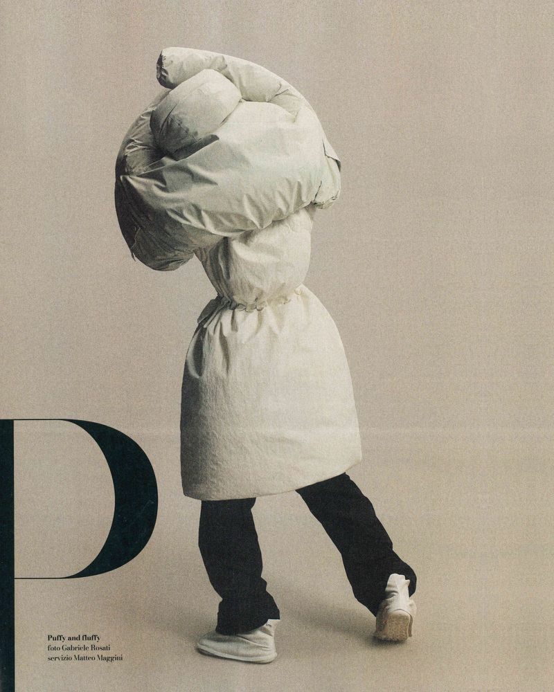 Fashionable person wearing a puffy coat covering head and torso, black pants, and white sneakers poses against a neutral background.
