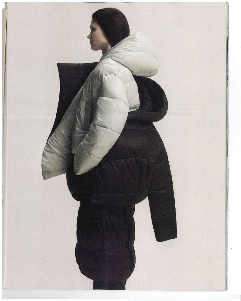 Fashion shot of a woman in a white hooded puffer jacket worn over a black puffer jacket against a pale backdrop in a magazine.