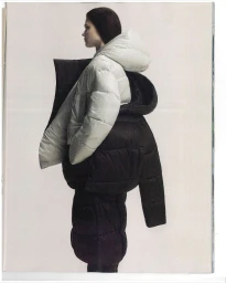Side profile of a model wearing layered white and black oversized sculptural puffer jackets against a plain white studio backdrop.