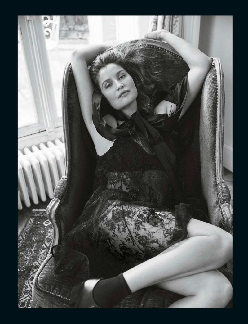 Elegant studio portrait of a brunette woman in a black lace dress lounging in a plush armchair, set against a light-filled window.