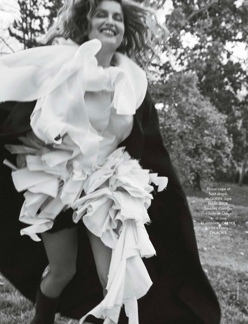 Fashion editorial style shot of a woman in a white frilly dress and cape running in a park setting with trees, captured at eye-level.