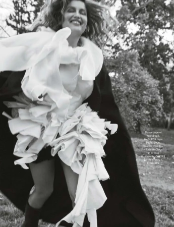 Fashion editorial style shot of a woman in a white frilly dress and cape running in a park setting with trees, captured at eye-level.