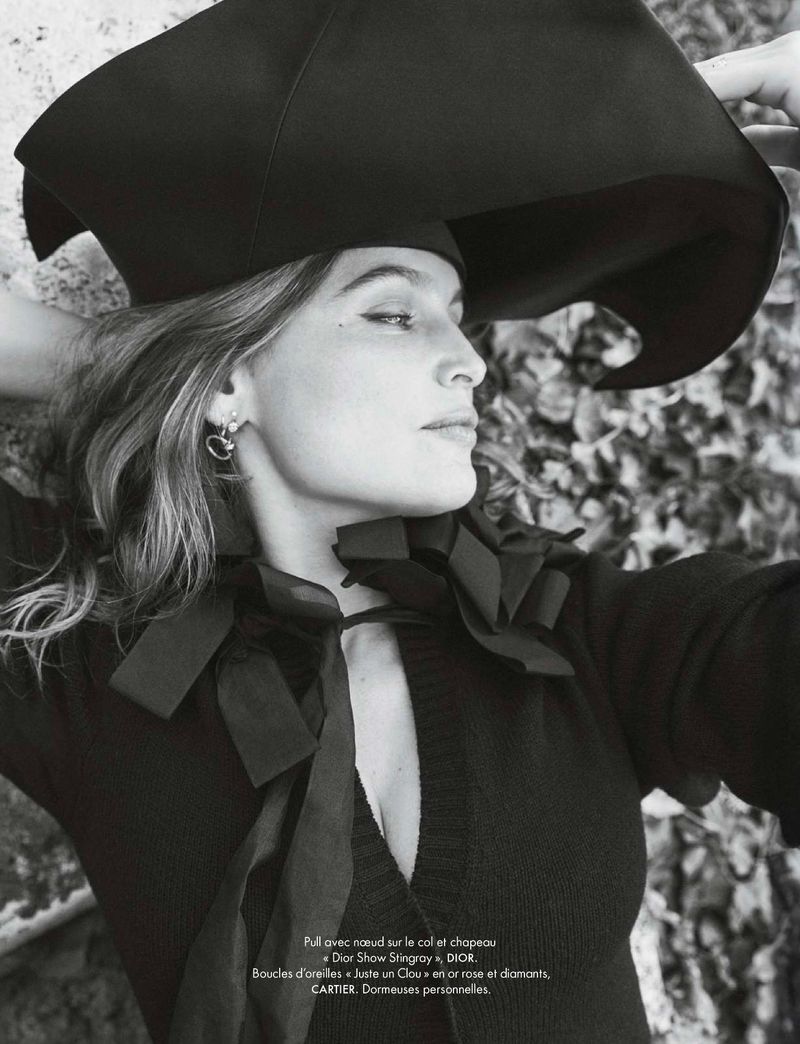 Monochrome close-up of a woman in black cardigan and floppy hat, wearing hoop earrings, against a leafy background in natural light.