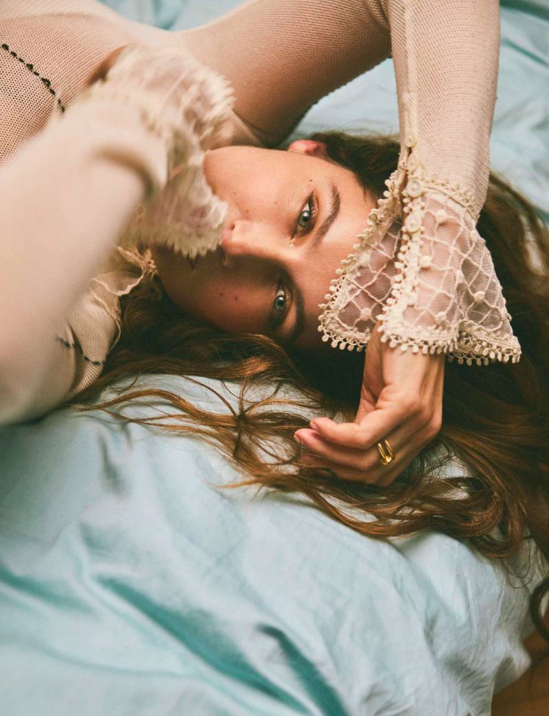Close-up portrait of a young adult female with green eyes laying on a teal-colored bed with long brown hair and a cream-colored lace top.