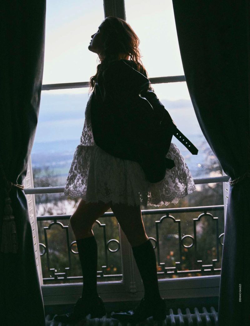 Silhouette of a woman in a white lace dress and boots, standing by a window overlooking a landscape, framed by curtains, bathed in backlight.