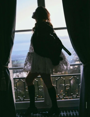 Silhouette of a woman in a white lace dress and boots, standing by a window overlooking a landscape, framed by curtains, bathed in backlight.
