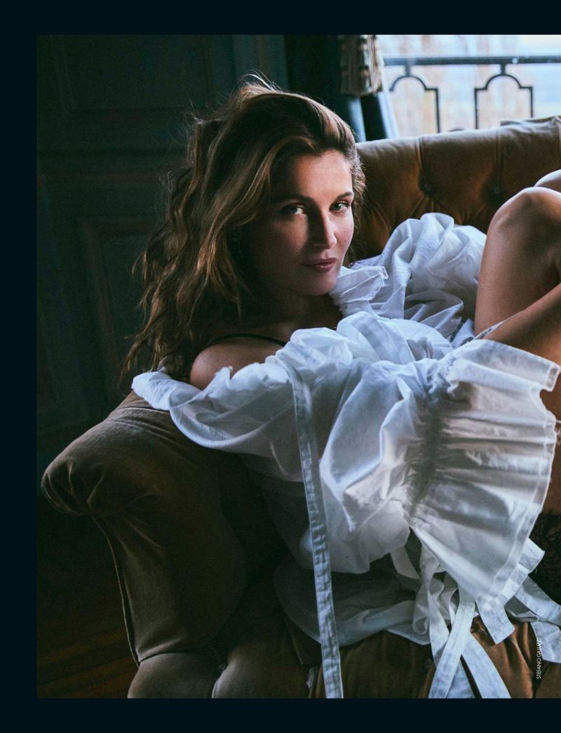 Laetitia Casta reclining on a brown tufted velvet sofa wearing a voluminous white ruffled off-the-shoulder blouse.