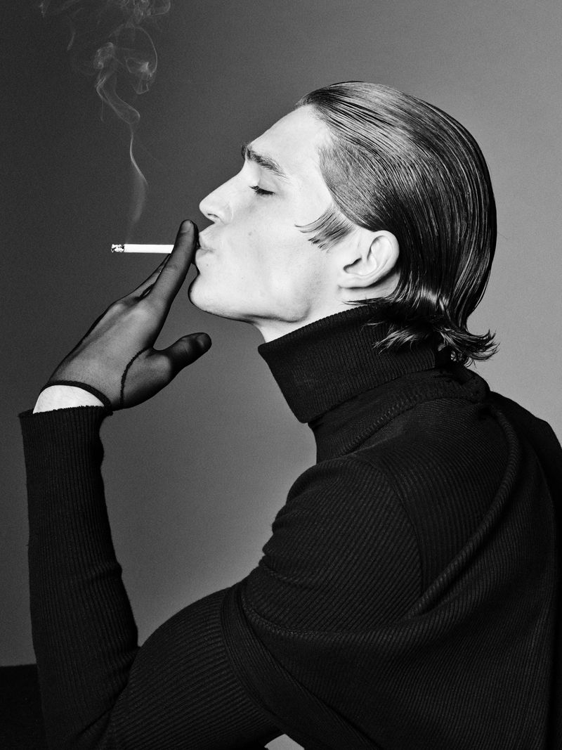 Monochrome portrait of a young adult man smoking a cigarette, wearing sheer gloves and a dark turtleneck sweater against a grey background.