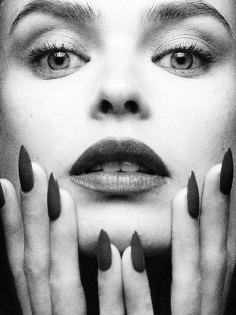 Monochrome portrait of a woman with dark lipstick and long nails holding hands to face in an indoor setting with studio lighting.