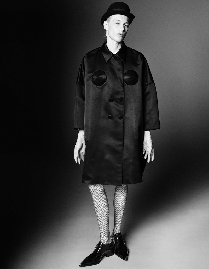 Black and white fashion portrait of an adult in a bowler hat, coat, fishnet stockings, and shoes against a dark gradient background.