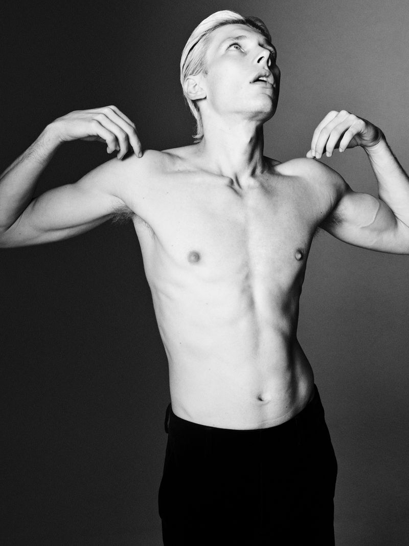Monochrome studio shot of shirtless young adult male with bleached hair, dark pants and raised arms looking upwards in a studio setting.