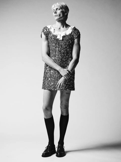 Black and white portrait of an adult male with short blonde hair wearing a sequined dress, knee socks, and shoes in a studio setting.
