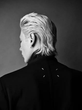 Back portrait of a platinum blond adult male with a black jacket, facing left against a neutral gray studio backdrop.